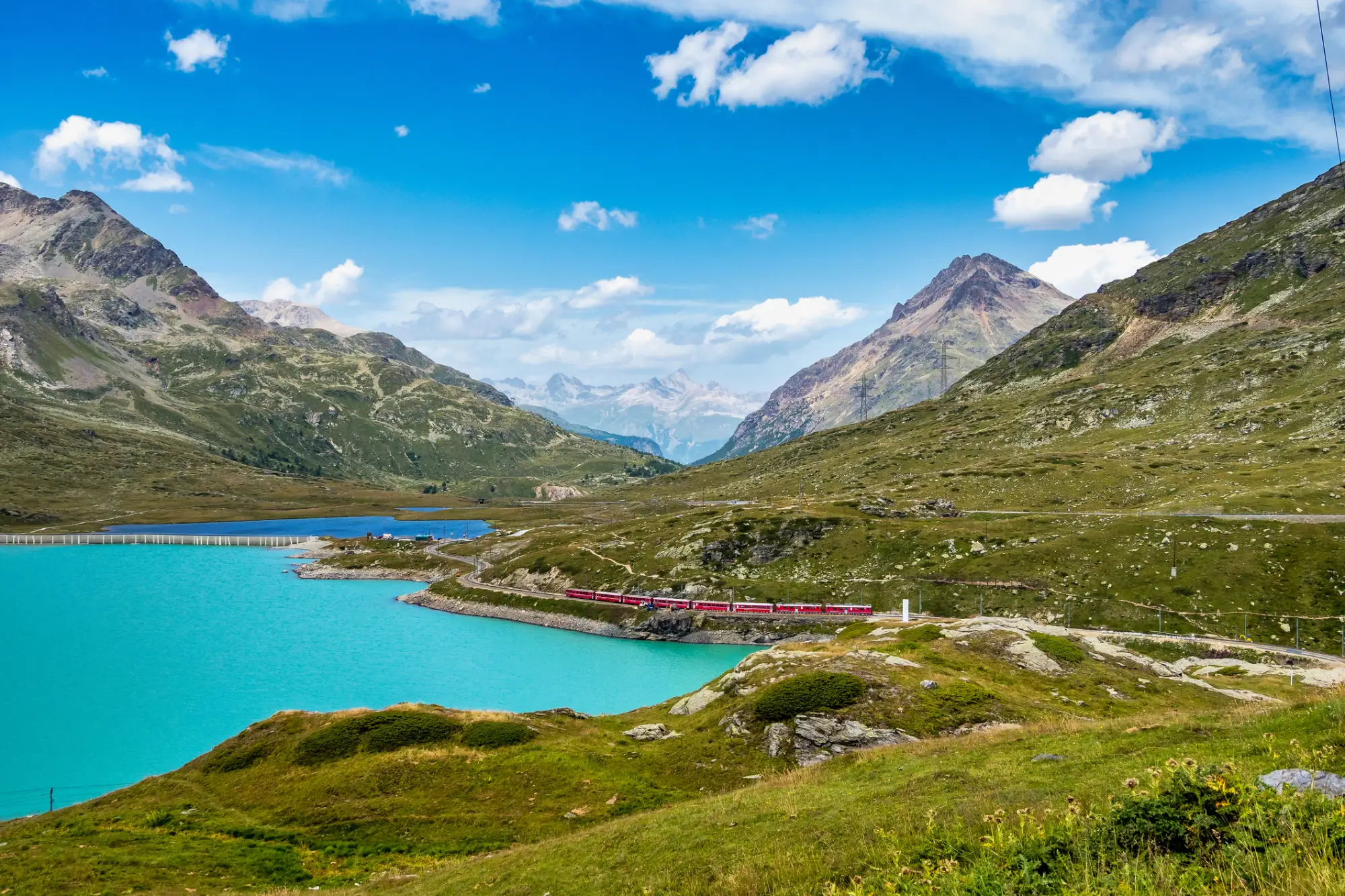 Rdeč vlak ob turkiznem gorskem jezeru v alpski pokrajini z gorami in modrim nebom v ozadju