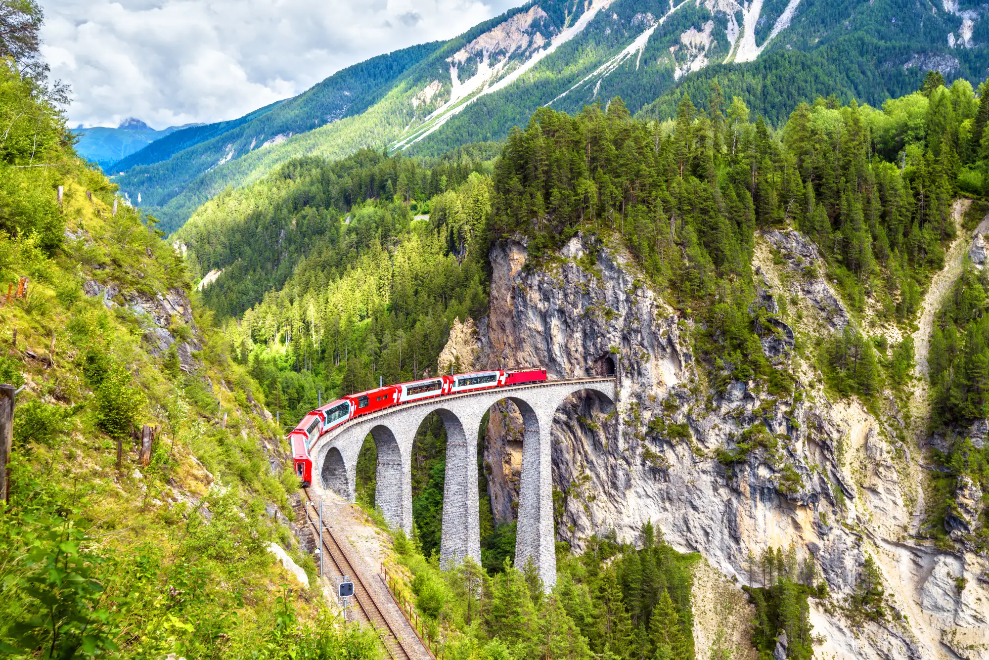 Rdeč panoramski vlak Bernina Express na kamnitem viaduktu skozi alpsko dolino z gozdovi in gorami v ozadju