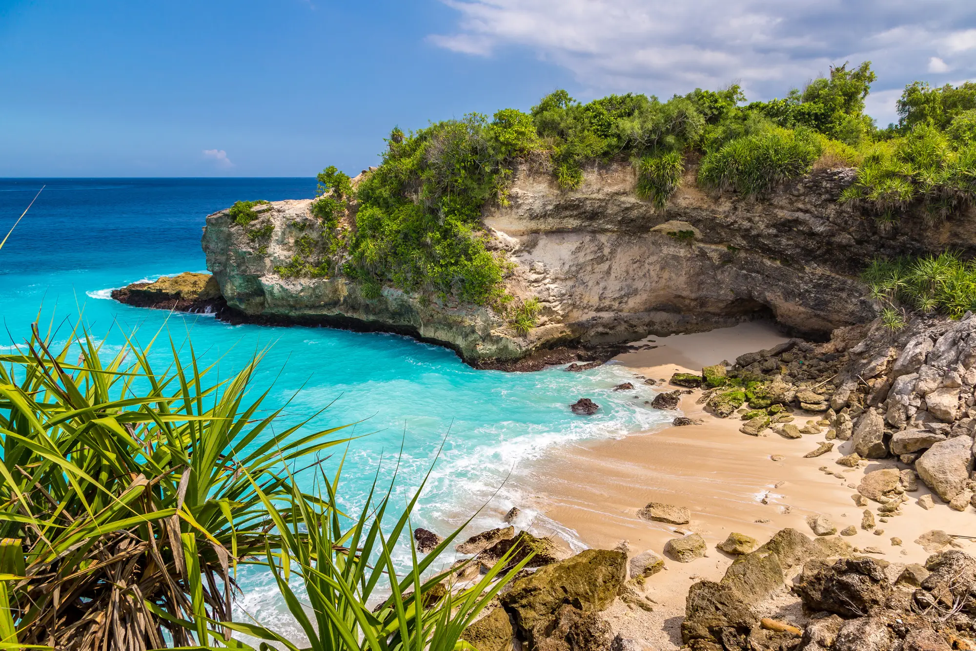 Pogled na Blue Lagoon Beach na Baliju z azurno modro vodo, peščeno plažo in tropsko naravo na skalah v ozadju
