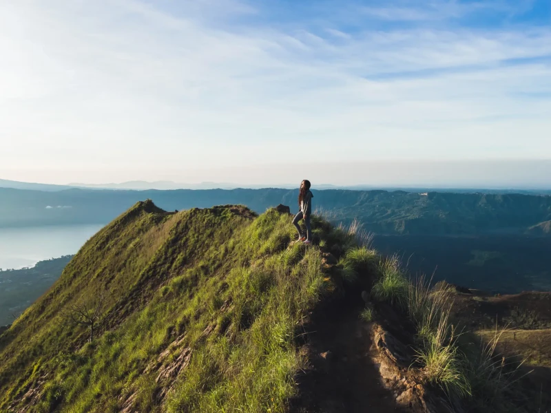 Popotnik stoji na grebenu vulkana Mt. Batur na Baliju ob sončnem vzhodu z razgledom na dolino in jezero.